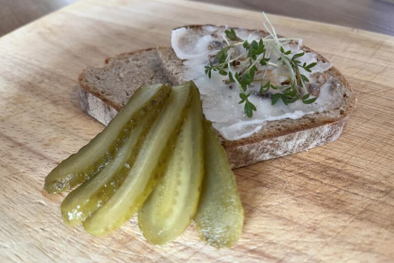 Apfel-Zwiebel-Schmalz auf Roggenbrot mit Kresse, daneben aufgefächerte Gewürzgurken auf einem Holzbrett